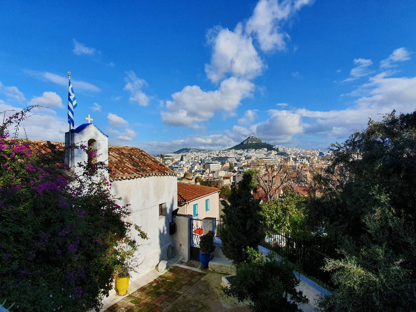 A whitewashed chapel with a blue-domed bell tower in Anafiotika, framed by bougainvillea, with Lycabettus Hill visible across the Athens rooftops