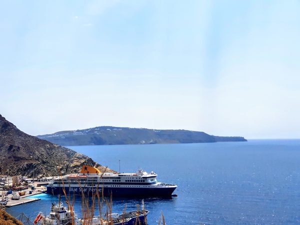 A Blue Star ferry docked at Santorini's Athinios port, with the switchback cliff road above and Thirasia island across the water