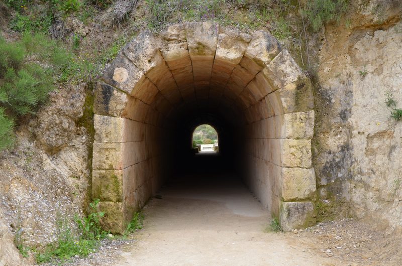 The ancient vaulted entrance tunnel of the Nemea stadium, a narrow stone passageway with arched ceiling leading from darkness toward daylight at the stadium end