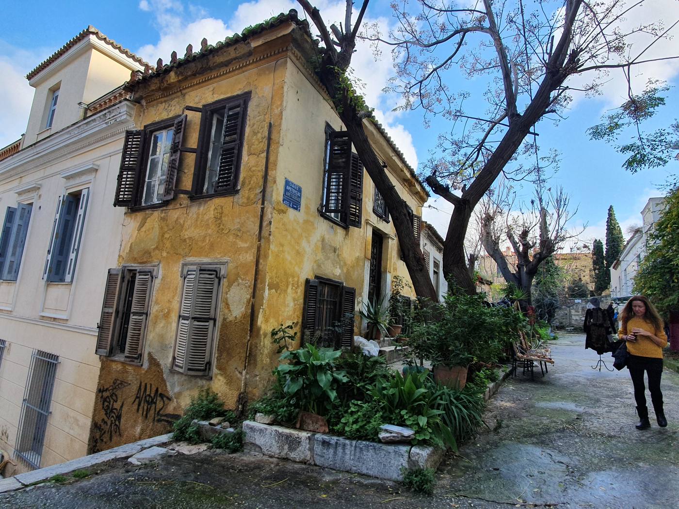 A quiet, ordinary street in the Plaka neighborhood with a pastel-colored old house, shuttered windows, and a pedestrian walking past