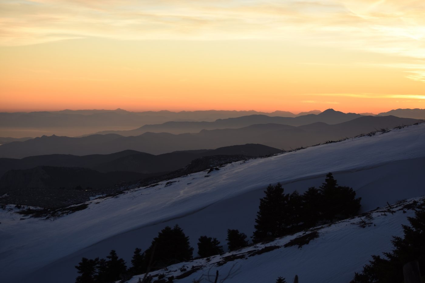 Sunset panorama from snow-covered Mount Parnitha slopes with layered mountain ridges and fir trees in the foreground