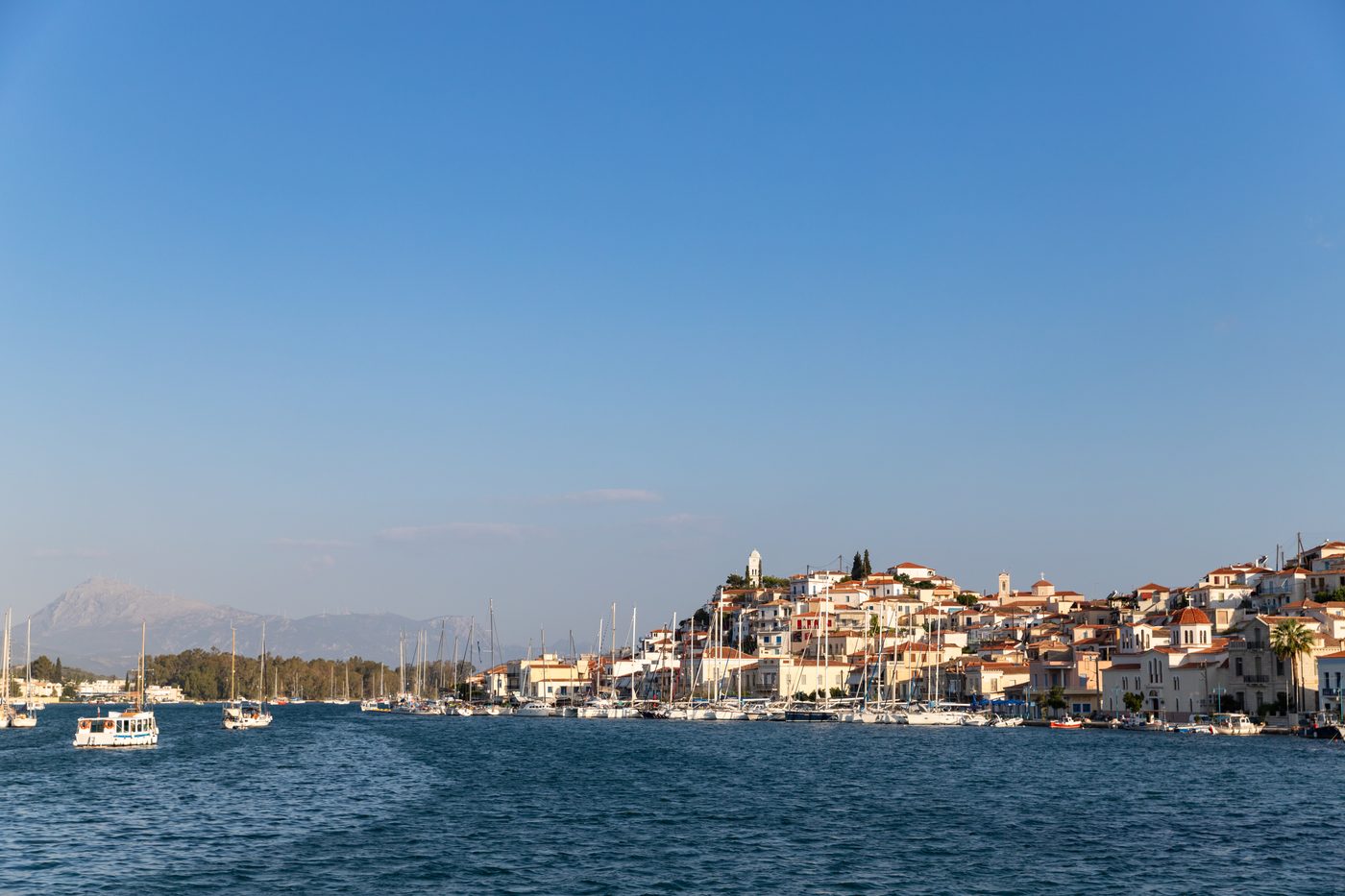 Poros town waterfront seen from the sea with white and terracotta-roofed buildings rising to the clock tower, yachts in the harbour and mountains beyond